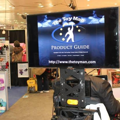 The Toy Man® in a power wheelchair with a 27 inch flat screen TV attached; displaying sponsors products at Toy Fair NY